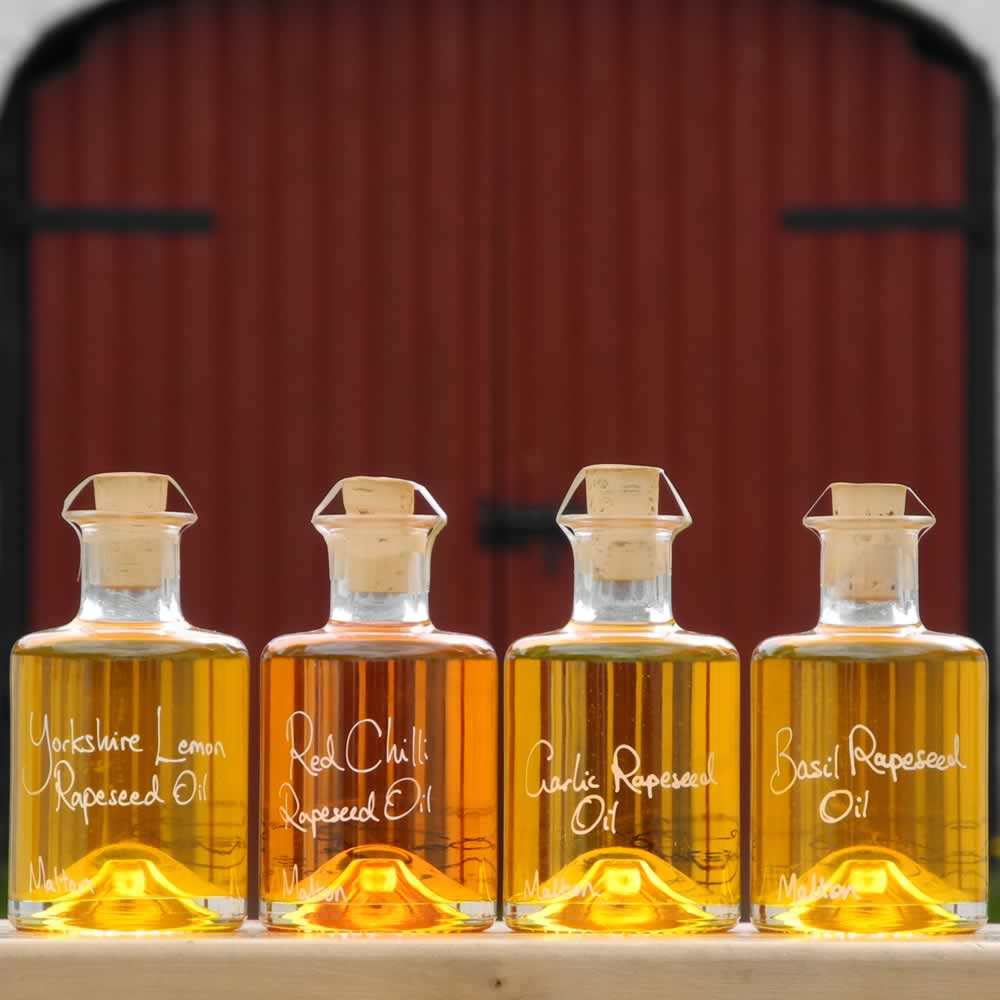 The Infused Oil Collection with Lemon Rapeseed Oil, Red Chilli Rapeseed Oil, Garlic Rapeseed Oil and Basil Rapeseed Oil in refillable 200ml bottles displayed in front of red barn doors