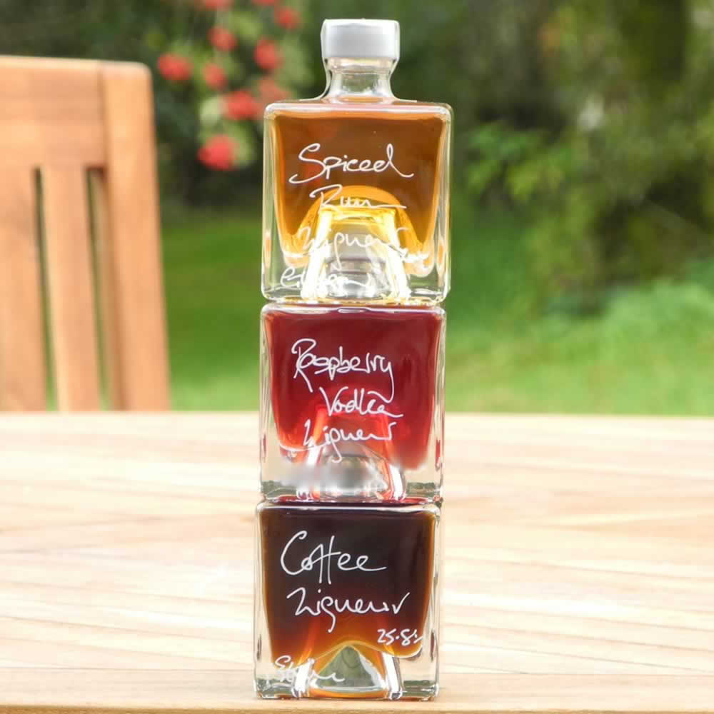 Three cube shaped 100ml stacking bottles of different handmade drinks, labeled by hand as Coffee Liqueur, Spiced Rum, and Raspberry Vodka, stacked on a table with a blurred background.