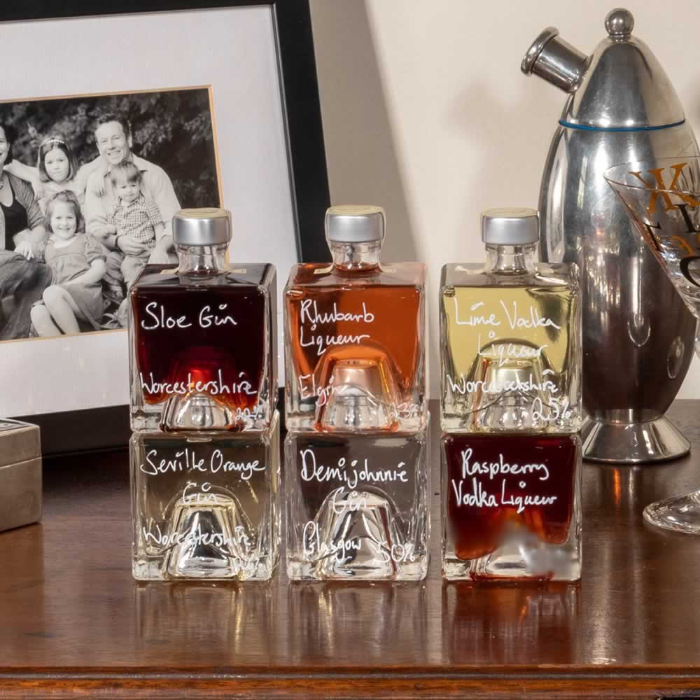 A set of six Mystic 100ml stacking glass bottles labeled with different types of handmade drinks from Sloe Gin to Raspberry Vodka, displayed on a wooden surface with a framed photograph and salad in the background.