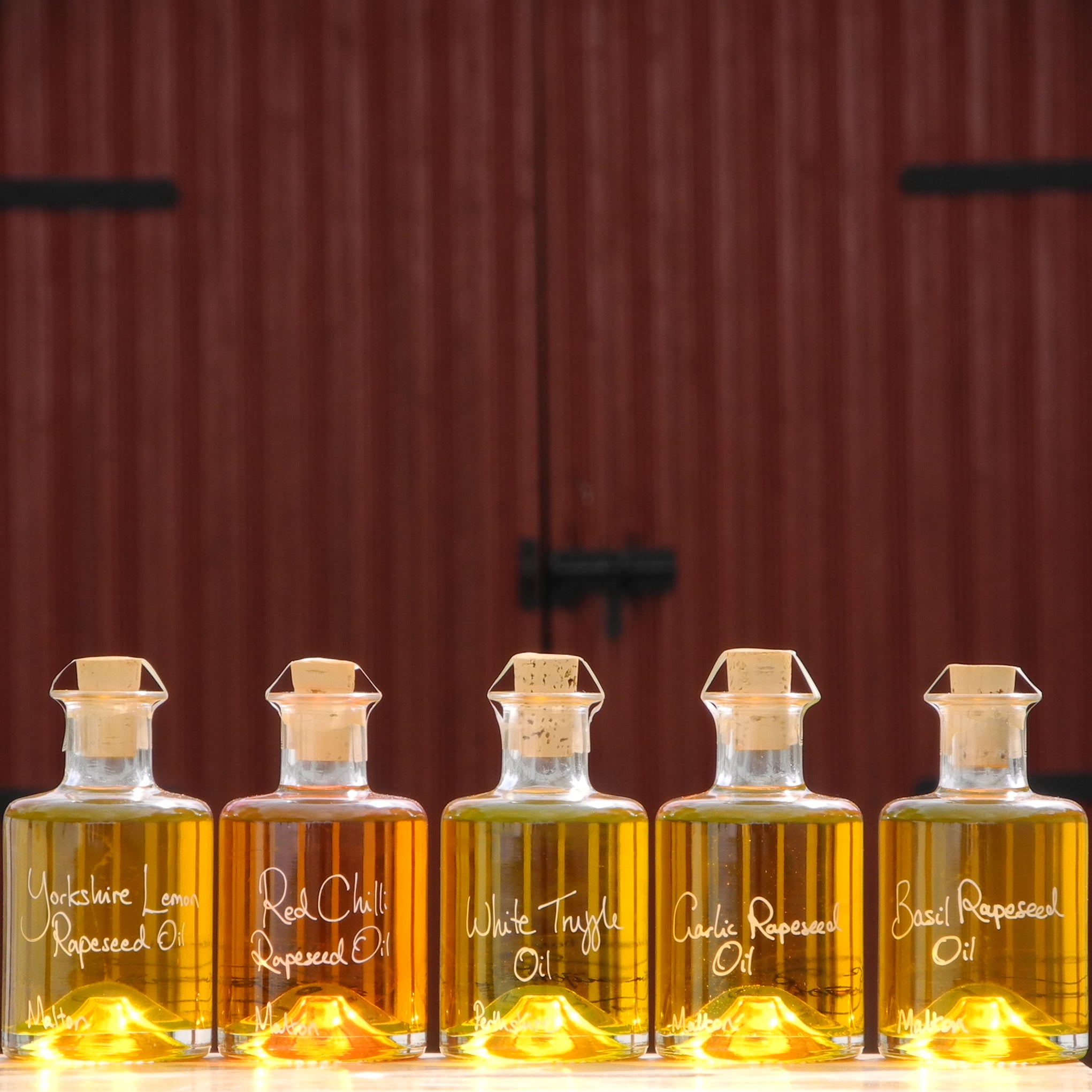 The Connoisseurs Infused Oil Collection with White Truffle Oil, Lemon Rapeseed Oil, Red Chilli Rapeseed Oil, Garlic Rapeseed Oil and Basil Rapeseed Oil in refillable 200ml bottles, this great fathers day gift is displayed in front of red barn doors. Personalised gift for cooks.