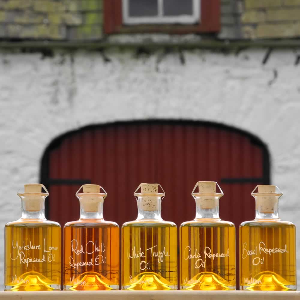 The Connoisseurs Infused Oil Collection with White Truffle Oil, Lemon Rapeseed Oil, Red Chilli Rapeseed Oil, Garlic Rapeseed Oil and Basil Rapeseed Oil in refillable 200ml bottles, this cooking gift set is displayed in front of red barn doors