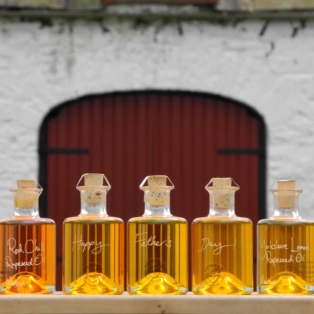 The Connoisseurs Infused Oil Collection with White Truffle Oil, Lemon Rapeseed Oil, Red Chilli Rapeseed Oil, Garlic Rapeseed Oil and Basil Rapeseed Oil in refillable 200ml bottles, this great fathers day gift is displayed in front of red barn doors. Personalised fathers day message written on bottles
