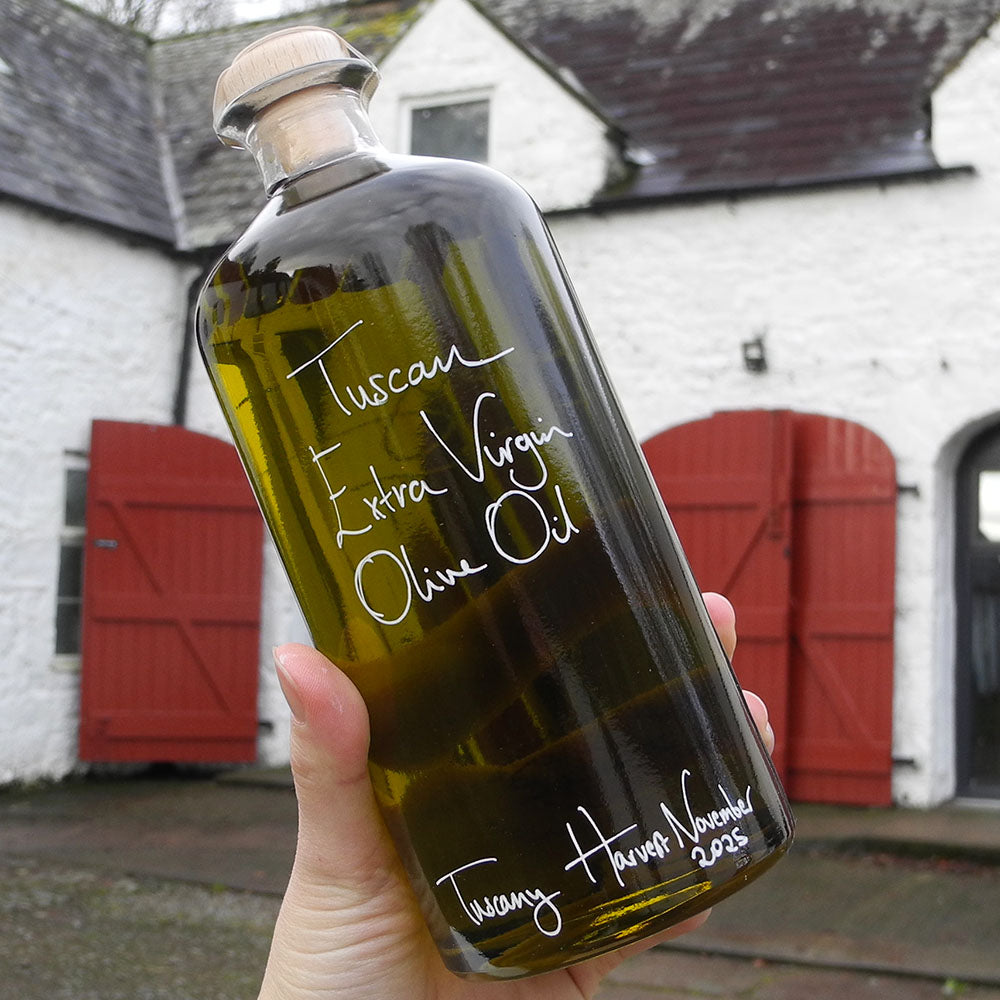 700ml bottle of Spinelli Tuscan Extra Virgin Olive Oil from Portugal with handwritten bottle message in white pen  being held in front of the red shutters at Glenlair Steading.