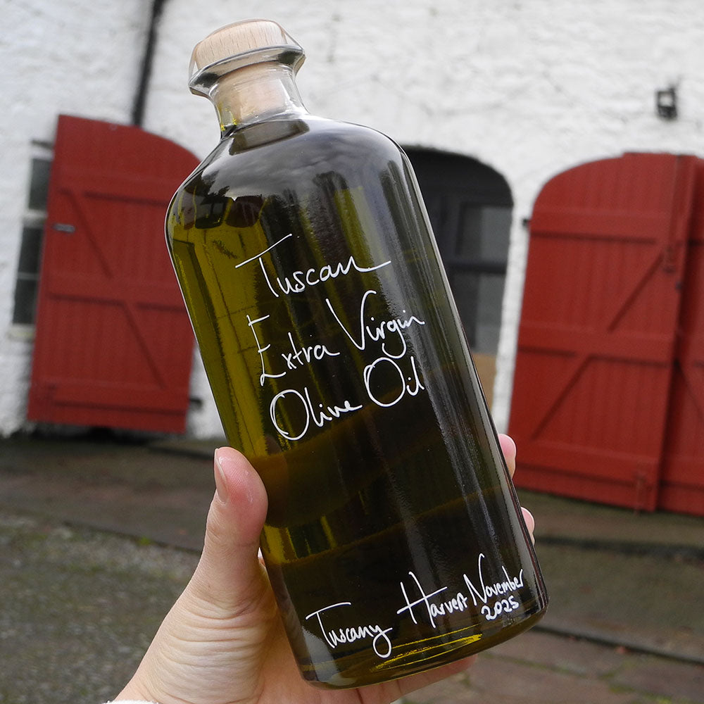 700ml bottle of Spinelli Tuscan Extra Virgin Olive Oil from Portugal with handwritten bottle message in white pen being held in front of the red shutters at Glenlair Steading.