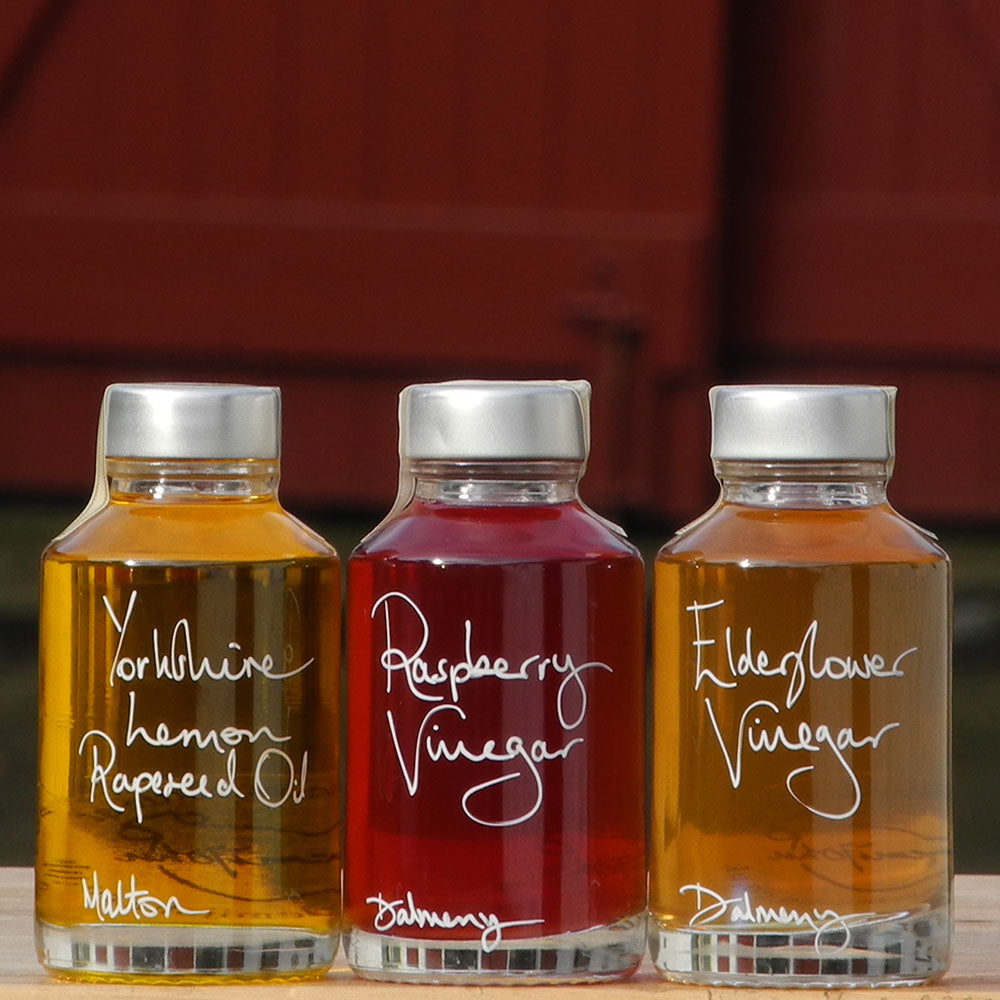The Mini Summer Salad Dressing Gift Set in three 100ml bottles containing Yorkshire Lemon Rapeseed Oil, Elderflower Vinegar and Raspberry Vinegar. The labels are handwritten in white pen in front of the red shutters at Glenlair Steading