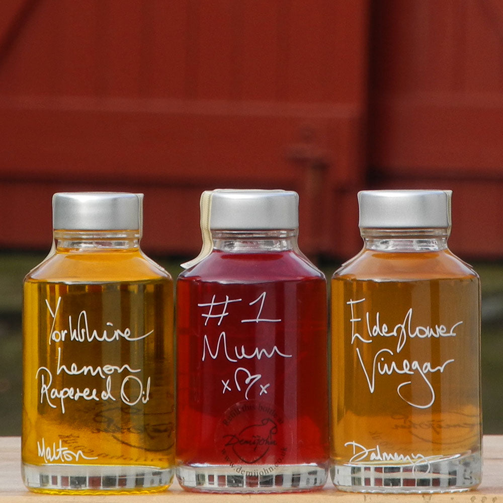 The Mini Summer Salad Dressing Gift Set in three 100ml bottles containing Yorkshire Lemon Rapeseed Oil, Elderflower Vinegar and Raspberry Vinegar. The labels are handwritten in white pen with a personalised message which says '#1 Mum' in front of the red shutters at Glenlair Steading