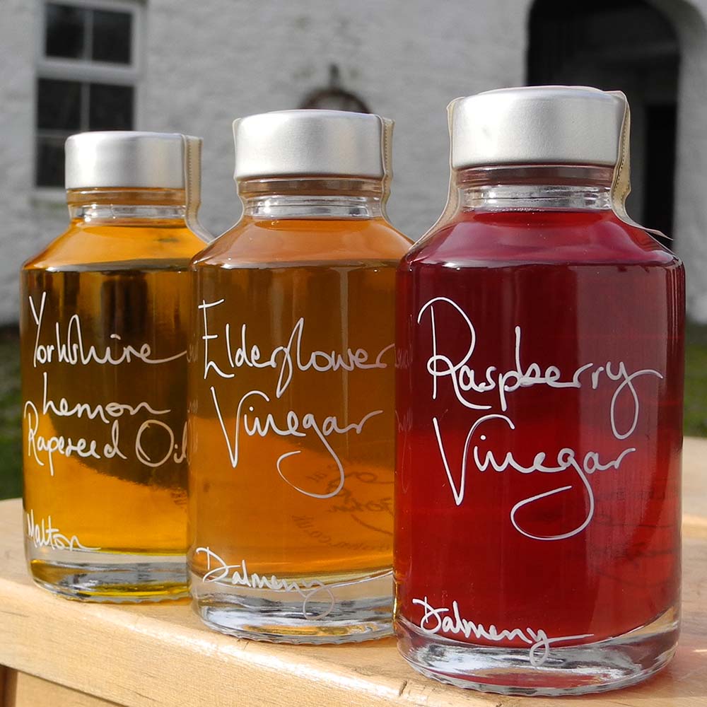 The Mini Summer Salad Dressing Gift Set in three 100ml bottles containing Yorkshire Lemon Rapeseed Oil, Elderflower Vinegar and Raspberry Vinegar. The labels are handwritten in white pen with a personalised message in front of the red shutters at Glenlair Steading