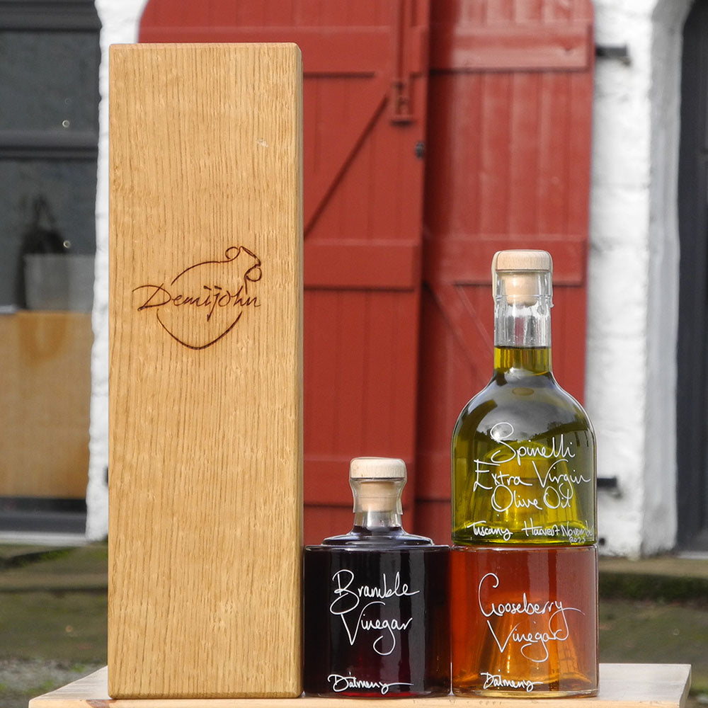 The Tuscan Oil and Vinegar Stacking Gift Set with Oak Gift Box where three clever stacking Impilabile 250ml bottles have been filled with fresh Spinelli Tuscan Extra Virgin Olive Oil, Gooseberry Vinegar and Bramble Vinegar, both from Edinburgh. The labels are handwritten in white pen in front of the red shutters at Glenlair Steading. The Oak Gift Box is next to the bottles with an engraved Demijohn logo on the front.