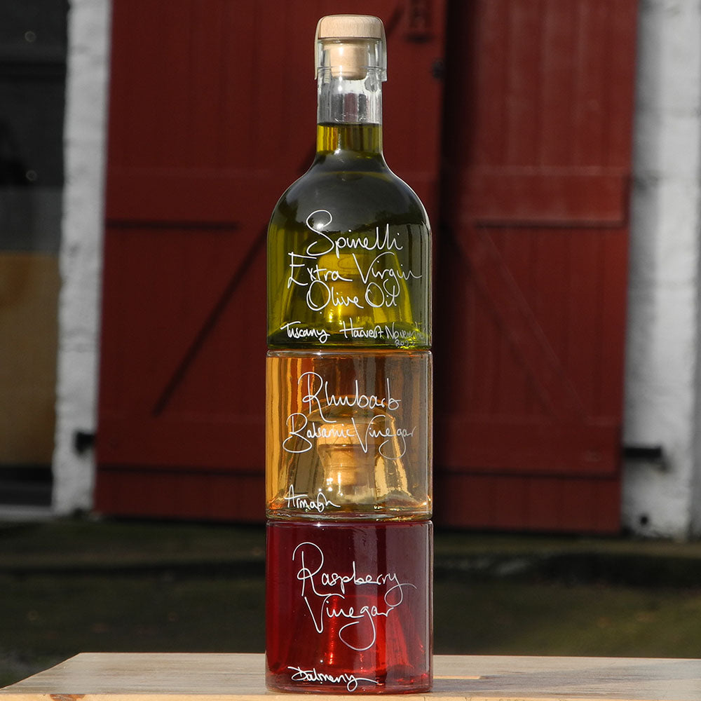 Tuscan Olive Oil and Vinegar Tower with three stacking bottles containing Spinelli Tuscan Extra Virgin Olive Oil, Rhubarb Balsamic Vinegar and Raspberry Vinegar, the labels are handwritten in white pen. The bottles are stacked in front of the red shutters at Glenlair Steading