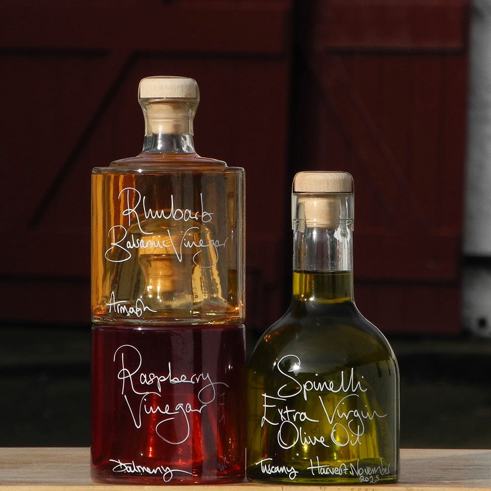 Tuscan Olive Oil and Vinegar Tower with three stacking bottles containing Spinelli Tuscan Extra Virgin Olive Oil, Rhubarb Balsamic Vinegar and Raspberry Vinegar, the labels are handwritten in white pen. The bottles are stacked in front of the red shutters at Glenlair Steading