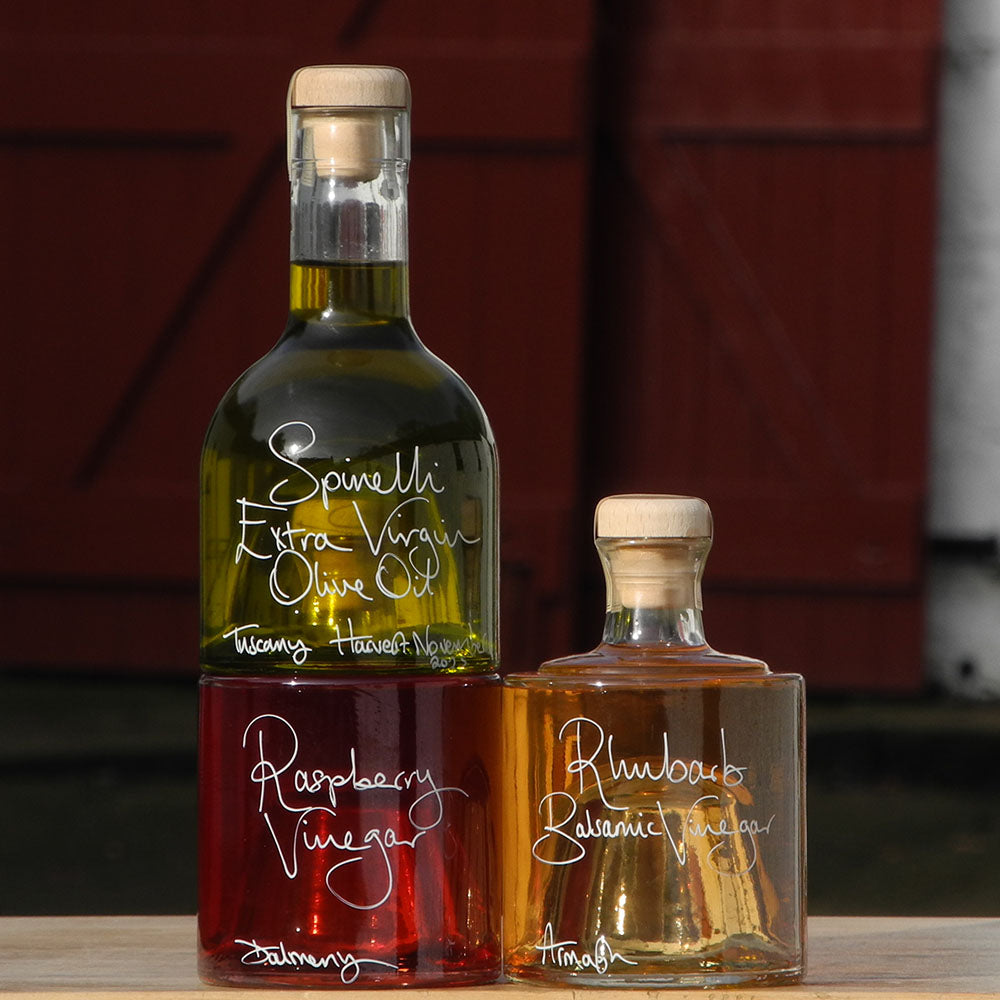 Tuscan Olive Oil and Vinegar Tower with three stacking bottles containing Spinelli Tuscan Extra Virgin Olive Oil, Rhubarb Balsamic Vinegar and Raspberry Vinegar, the labels are handwritten in white pen. The bottles are stacked in front of the red shutters at Glenlair Steading