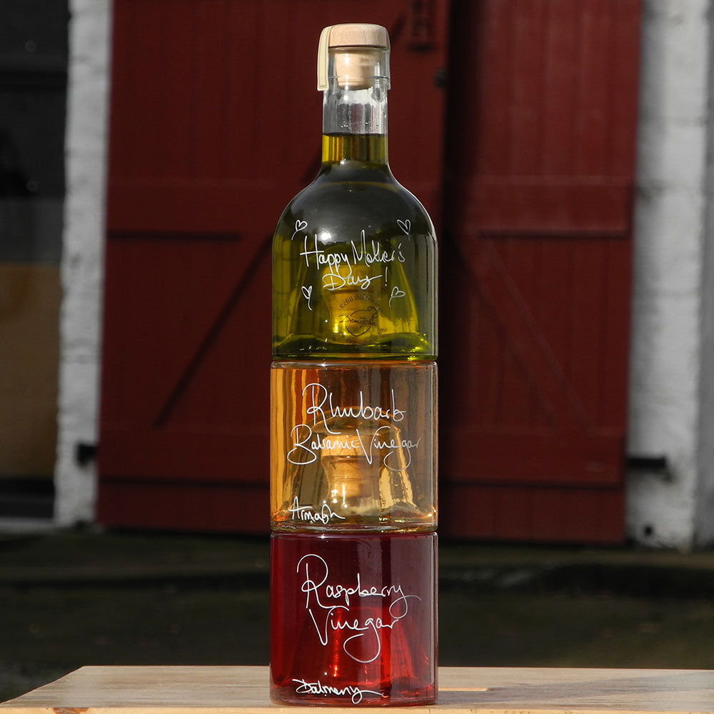 Tuscan Olive Oil and Vinegar Tower with three stacking bottles containing Spinelli Tuscan Extra Virgin Olive Oil, Rhubarb Balsamic Vinegar and Raspberry Vinegar, the labels are handwritten in white pen with personalised message that says Happy Mother's Day. The bottles are stacked in front of the red shutters at Glenlair Steading
