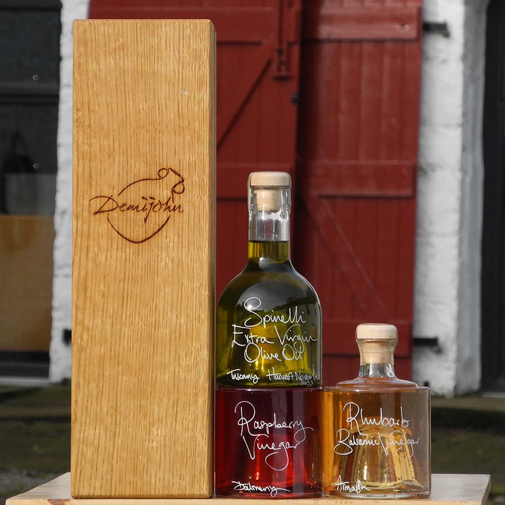 Tuscan Olive Oil and Vinegar Tower with Oak Gift Box has three stacking bottles containing Spinelli Tuscan Extra Virgin Olive Oil, Rhubarb Balsamic Vinegar and Raspberry Vinegar, the labels are handwritten in white pen. The bottles are stacked in front of the red shutters at Glenlair Steading