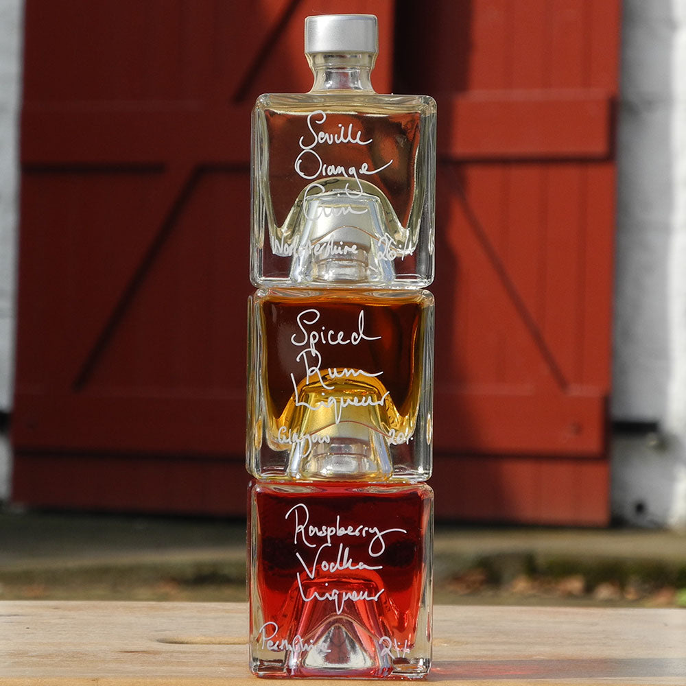 A gift set of three stacked 100ml cube shaped glass bottles with handmade drinks, each labeled with a different type of liqueur, against a red barn door background.