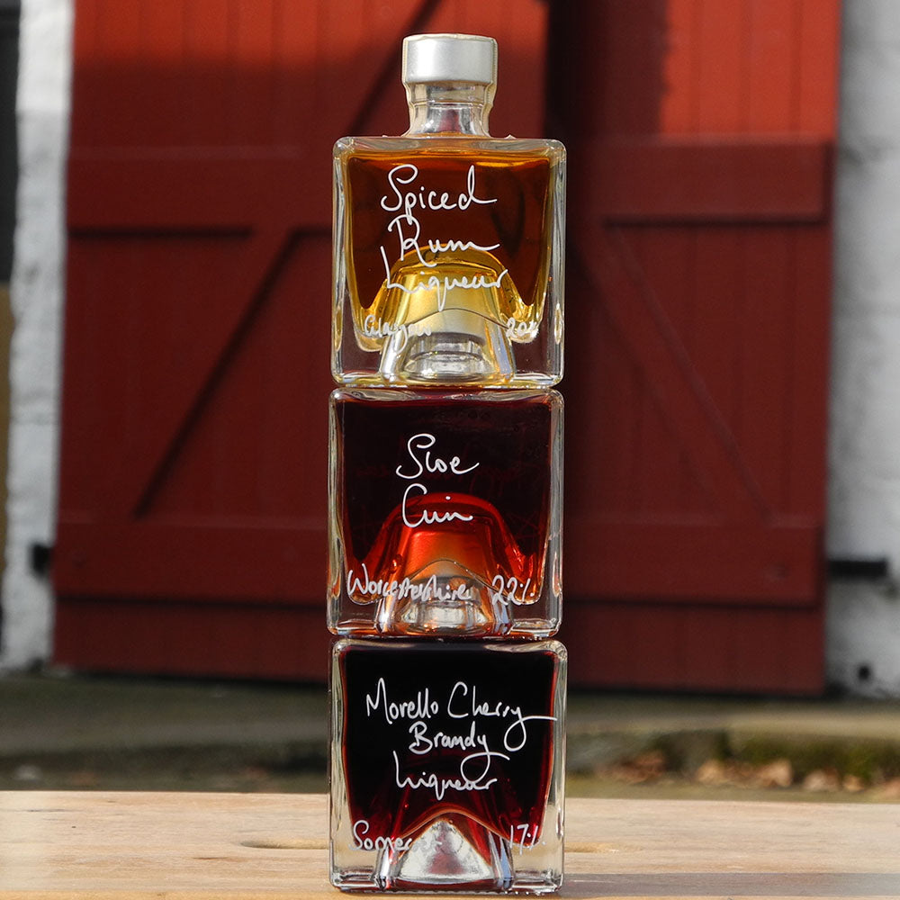 Three cube stacking bottles are filled with Spiced Rum Liqueur, Sloe Gin and Morello Cherry Brandy Liqueur handwritten in white pen in front of the red shutters of Demijohn at Glenlair Steading.