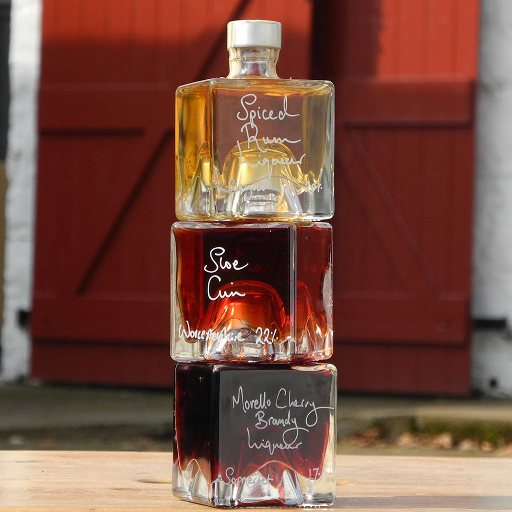 Three cube stacking bottles are filled with Spiced Rum Liqueur, Sloe Gin and Morello Cherry Brandy Liqueur handwritten in white pen in front of the red shutters of Demijohn at Glenlair Steading.