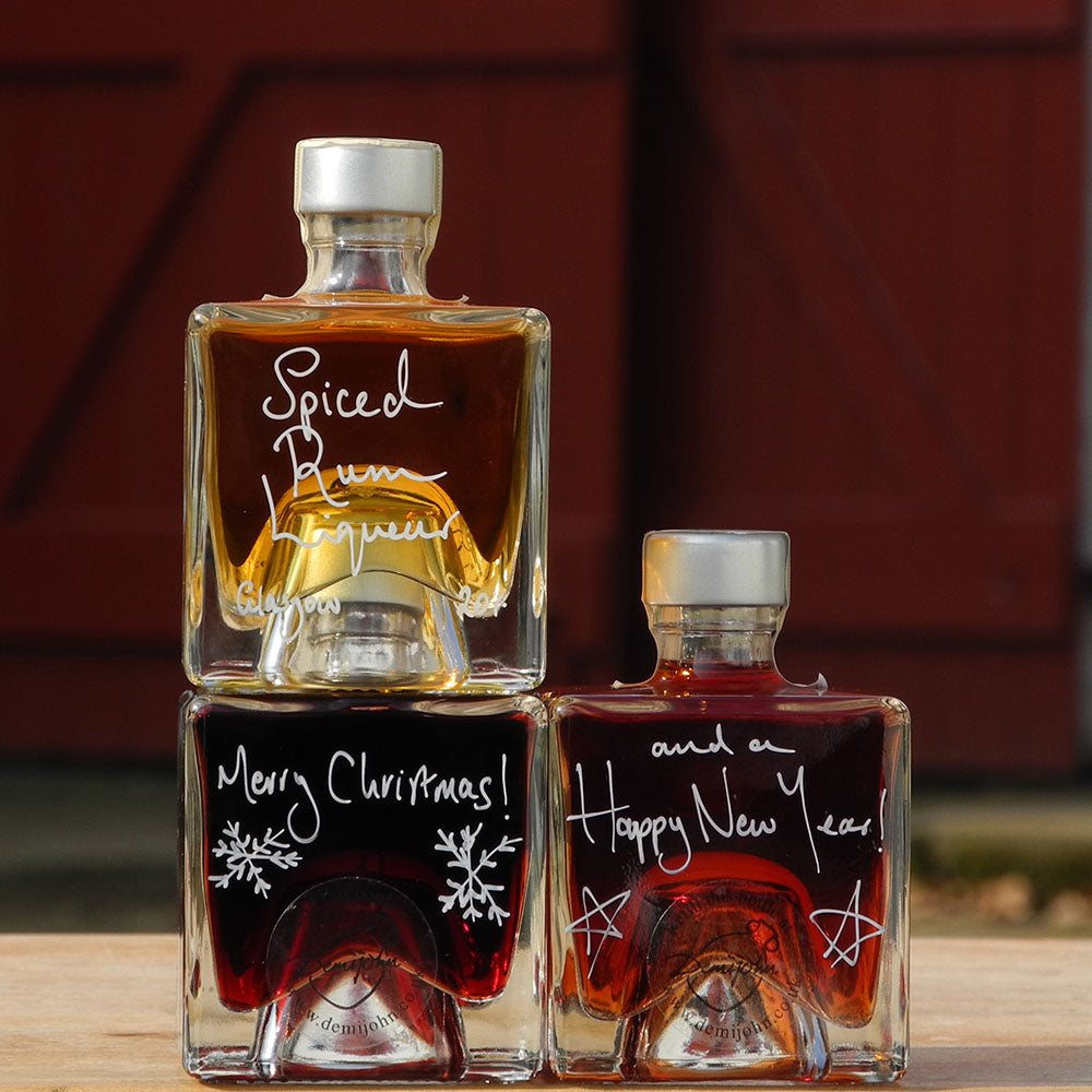 Three cube stacking bottles are filled with Spiced Rum Liqueur, Sloe Gin and Morello Cherry Brandy Liqueur handwritten in white pen in front of the red shutters of Demijohn at Glenlair Steading. There is a personalised message which says Merry Christmas and a Happy New Year spread over a couple of the bottles.
