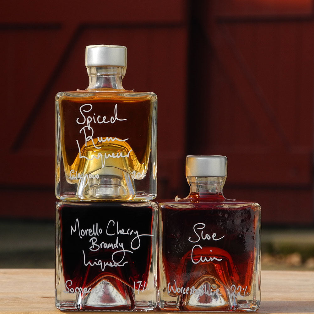 Three cube stacking bottles are filled with Spiced Rum Liqueur, Sloe Gin and Morello Cherry Brandy Liqueur handwritten in white pen in front of the red shutters of Demijohn at Glenlair Steading. 
