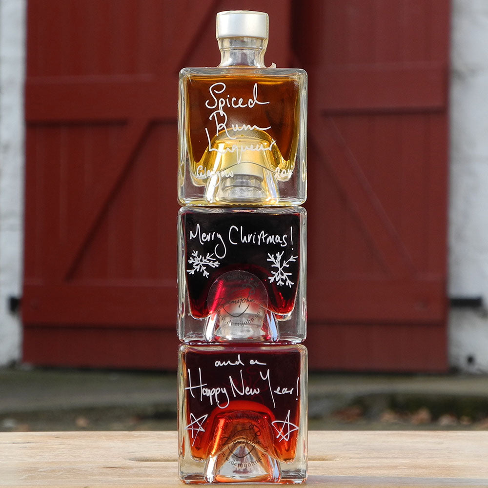 Three cube stacking bottles are filled with Spiced Rum Liqueur, Sloe Gin and Morello Cherry Brandy Liqueur handwritten in white pen in front of the red shutters of Demijohn at Glenlair Steading. There is a personalised message which says Merry Christmas and a Happy New Year spread over a couple of the bottles.