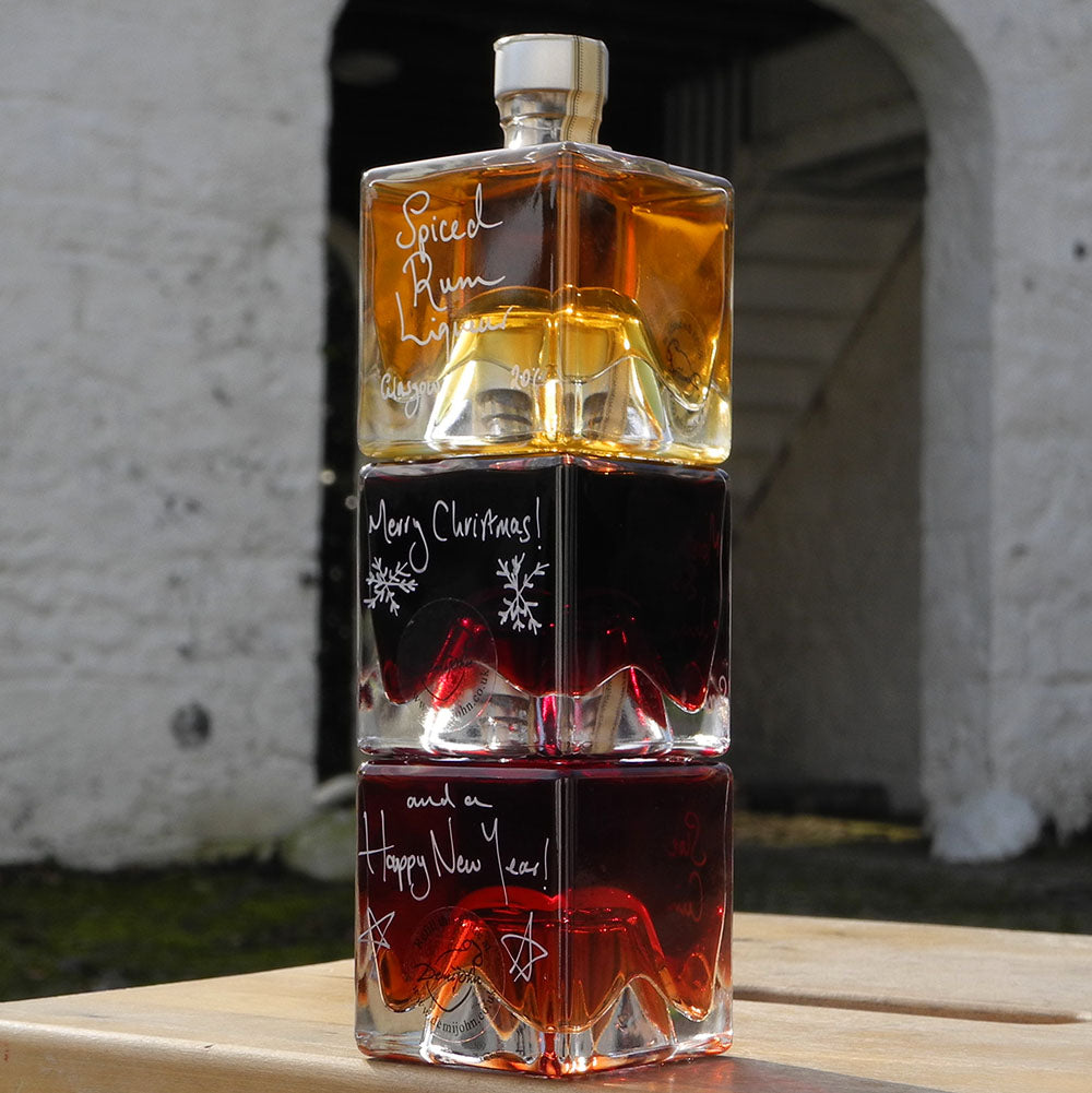 Three cube stacking bottles are filled with Spiced Rum Liqueur, Sloe Gin and Morello Cherry Brandy Liqueur handwritten in white pen in front of the white walls of Demijohn at Glenlair Steading. There is a personalised message which says Merry Christmas and a Happy New Year spread over a couple of the bottles.