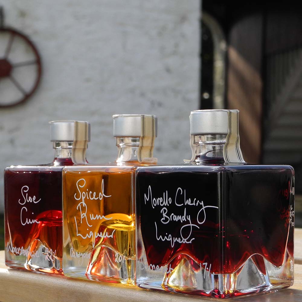 Three cube stacking bottles are filled with Spiced Rum Liqueur, Sloe Gin and Morello Cherry Brandy Liqueur handwritten in white pen in front of the white walls of Demijohn at Glenlair Steading.