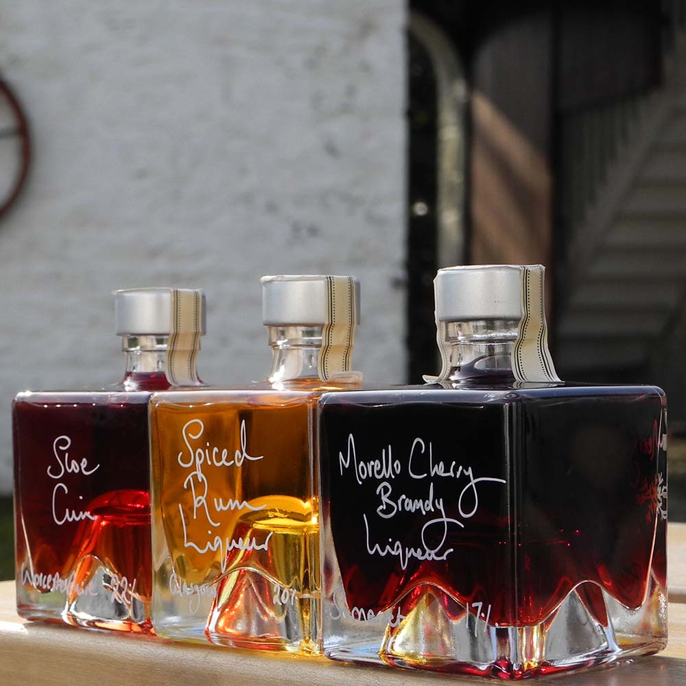 Three cube stacking bottles are filled with Spiced Rum Liqueur, Sloe Gin and Morello Cherry Brandy Liqueur handwritten in white pen in front of the red shutters of Demijohn at Glenlair Steading. 
