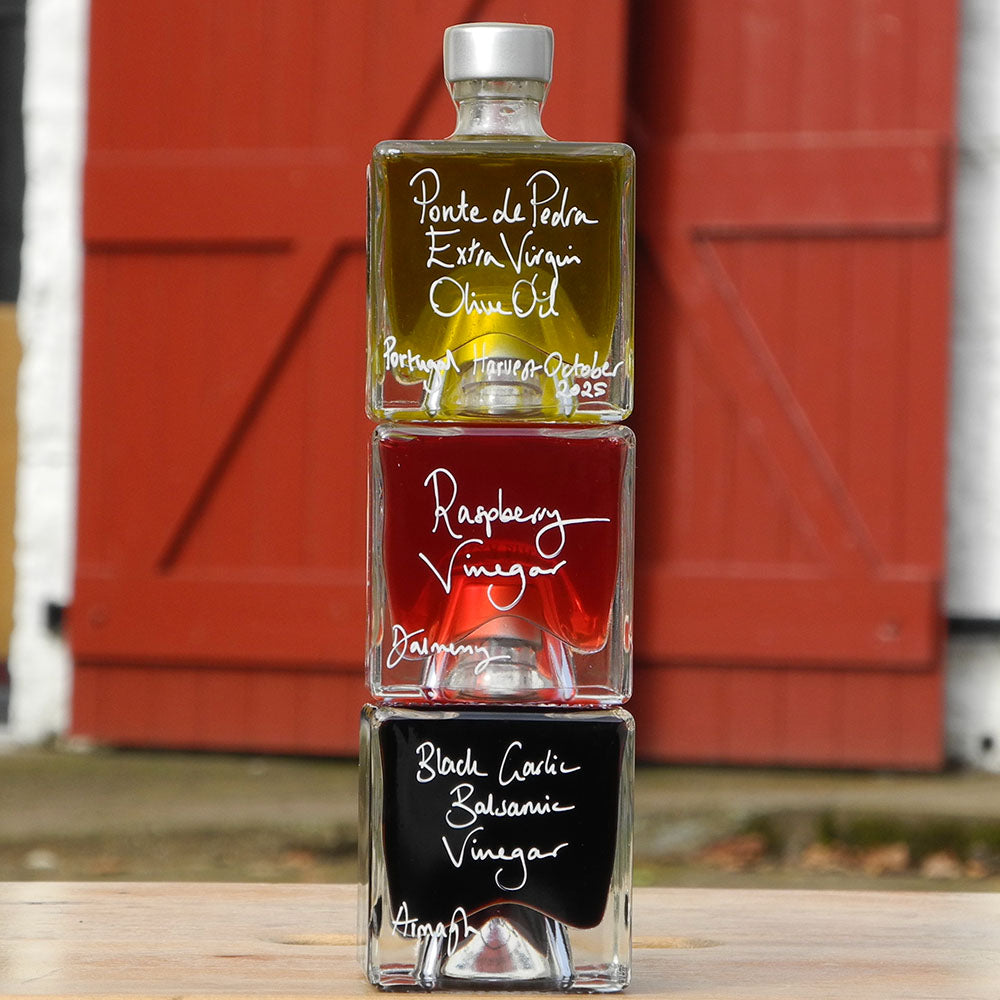 Three stacked square glass bottles of vinegar on a wooden surface in front of a red barn door. The top bottle contains olive oil and is labeled 'Ponte de Pedra Extra Virgin Olive Oil'; the middle bottle contains raspberry vinegar and is labeled 'Raspberry Vinegar'; the bottom bottle contains black garlic balsamic vinegar and is labeled 'Black Garlic Balsamic Vinegar'.
