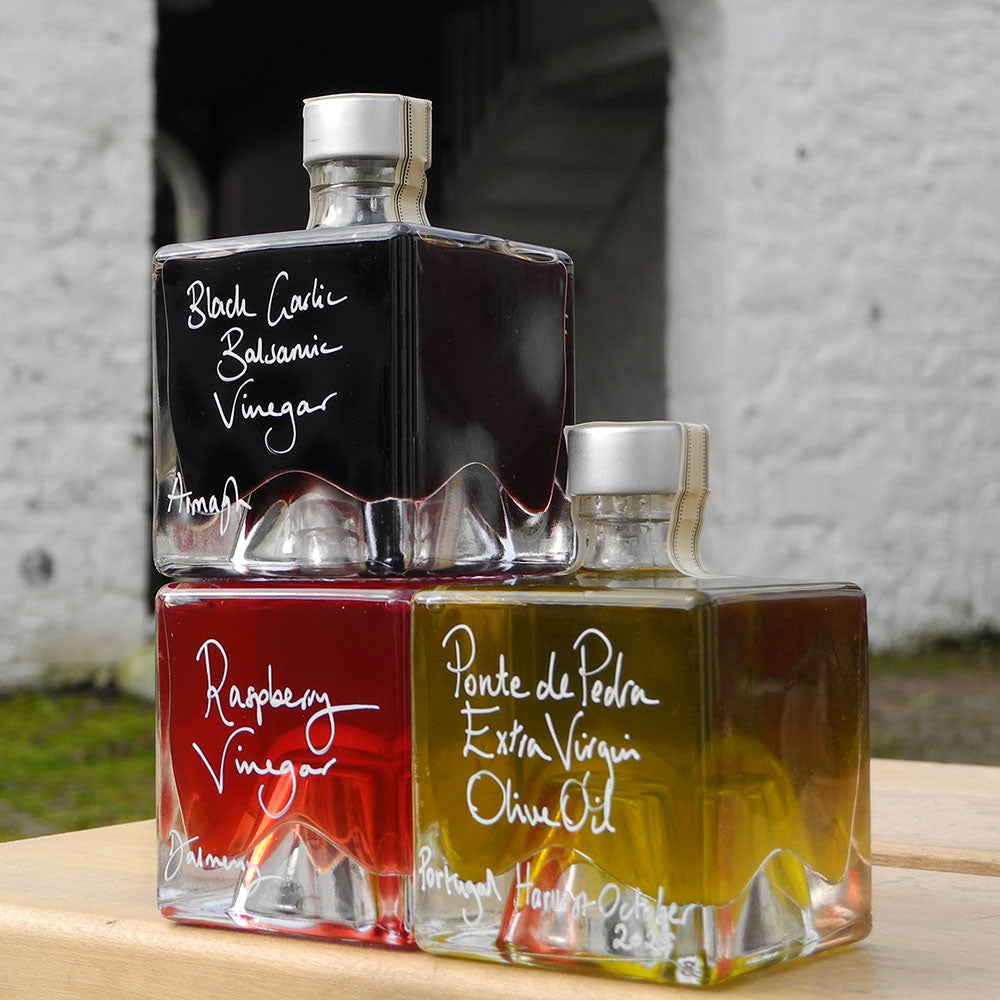 Three square glass bottles of vinegar and olive oil stacked on a wooden surface outdoors. The top bottle contains black carvi balsamic vinegar, the bottom left contains raspberry vinegar, and the bottom right contains ponte de oliva extra virgin olive oil.