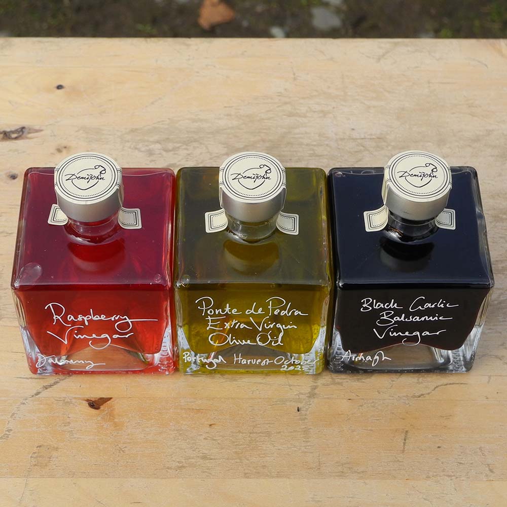 Three 100ml square glass bottles lined up on a wooden surface. The bottles contain red raspberry vinegar, Ponte de Pedra Extra Virgin Oil and Black Garlic Balsamic Vinegar, each with a silver cap and white handwritten text labels.