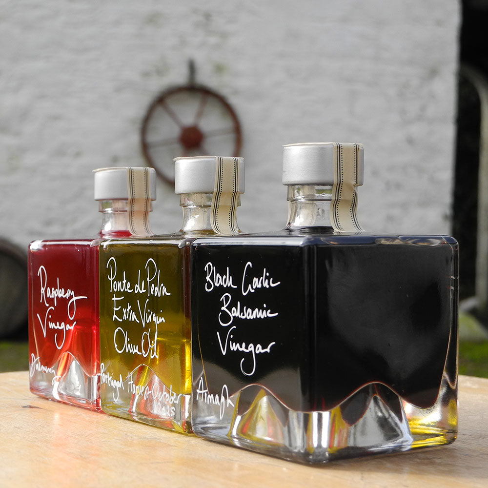 Three 100ml square glass bottles lined up on a wooden surface. The bottles contain red raspberry vinegar, Ponte de Pedra Extra Virgin Oil and Black Garlic Balsamic Vinegar, each with a silver cap and white handwritten text labels.