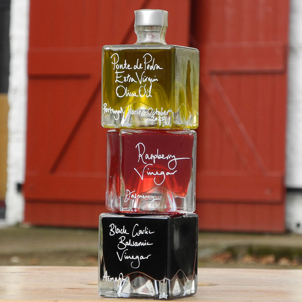 Three 100ml square glass bottles stacked up as a gift set on a wooden surface. The bottles contain Scottish Raspberry Vinegar, Ponte de Pedra Extra Virgin Oil and Black Garlic Balsamic Vinegar, each with a silver cap and white handwritten text labels.