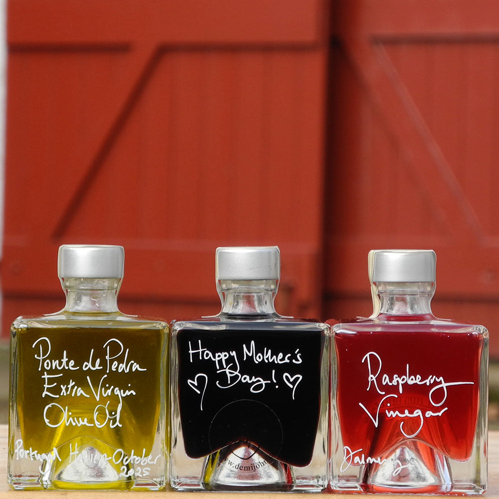 Three 100ml square glass bottles on a wooden surface. The bottles contain Scottish Raspberry Vinegar, Ponte de Pedra Extra Virgin Oil and Black Garlic Balsamic Vinegar, each with a silver cap and white handwritten text labels. The Balsamic Vinegar has a handwritten mesage saying Happy Mother's Day on it.