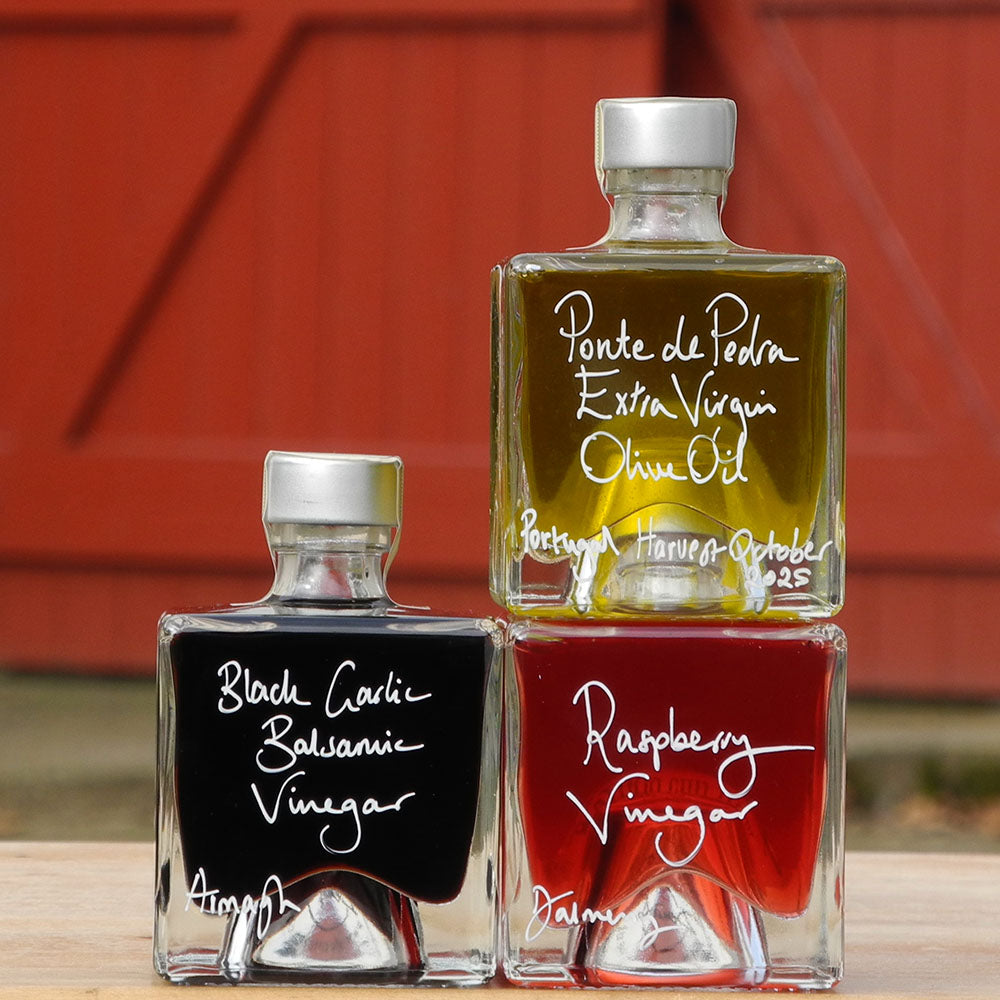 Three 100ml square glass bottles on a wooden surface. The bottles contain Scottish Raspberry Vinegar, Ponte de Pedra Extra Virgin Oil and Black Garlic Balsamic Vinegar, each with a silver cap and white handwritten text labels.