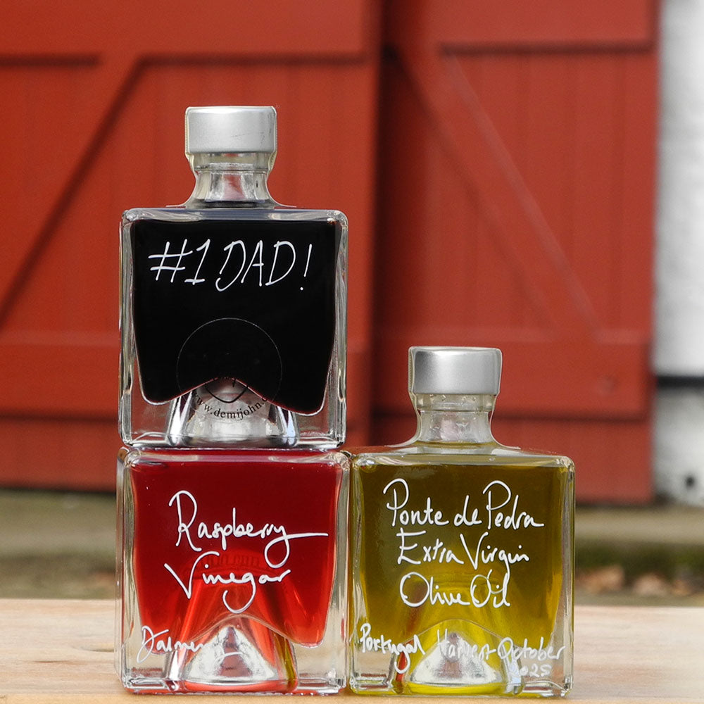 Three 100ml square glass bottles on a wooden surface. The bottles contain Scottish Raspberry Vinegar, Ponte de Pedra Extra Virgin Oil and Black Garlic Balsamic Vinegar, each with a silver cap and white handwritten text labels. The Balsamic Vinegar has a handwritten mesage saying #1 Dad on it.