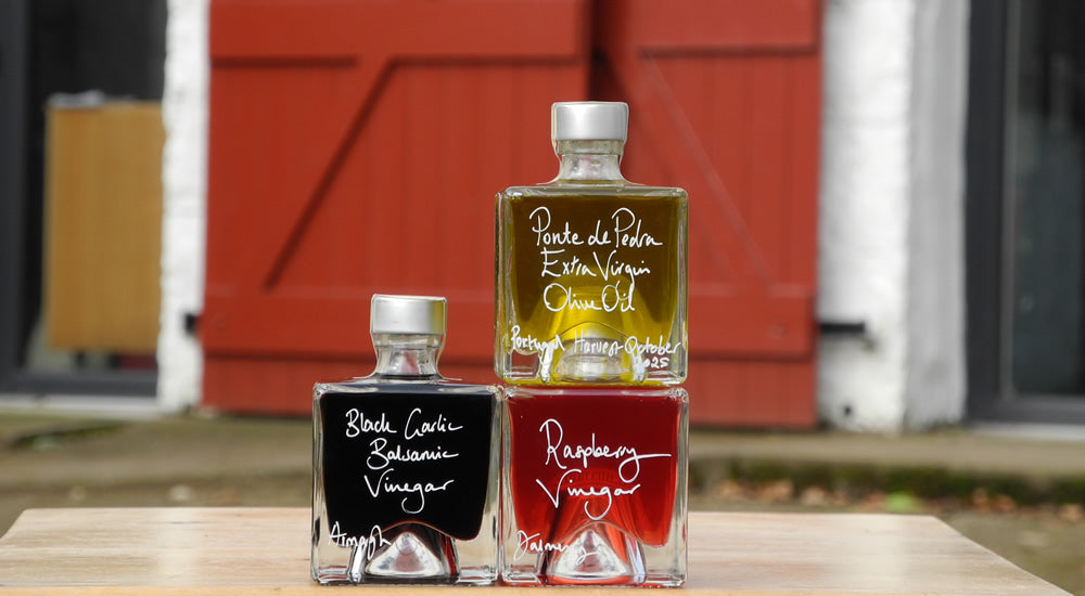 Three square glass bottles of vinegar arranged on a wooden surface in front of a red barn door. The bottles contain black, red, and golden-yellow liquids, with white handwritten labels reading 'Black Garlic Balsamic Vinegar,' 'Ponte de Pedra Extra Vergin Olive Oil,' and 'Raspberry Vinegar.
