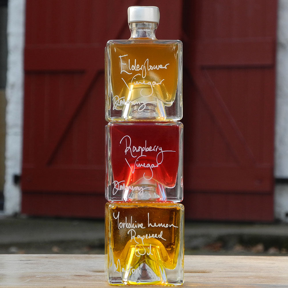 Three stacking, cube shaped 100ml glass bottles of Elderflower Vinegar, Scottish Raspberry Vinegar and Lemon infused Rapseed Oil, photographed outdoors in front of red barn doors.