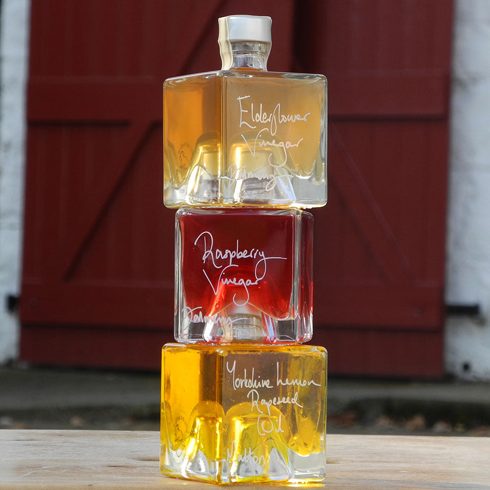 Three stacking, cube shaped, refillable 100ml glass bottles of Elderflower Vinegar, Scottish Raspberry Vinegar and Lemon infused Rapseed Oil, photographed outdoors in front of red barn doors.