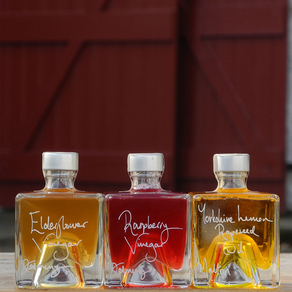 Three cube shaped, refillable 100ml glass bottles of Elderflower Vinegar, Scottish Raspberry Vinegar and Lemon infused Rapseed Oil, photographed outdoors in front of red barn doors.