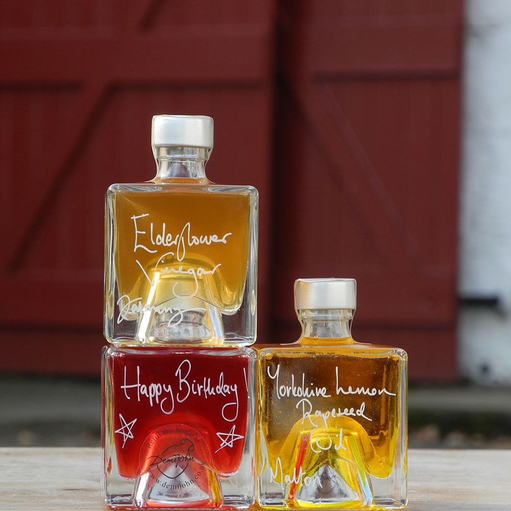 Three stacking, cube shaped, refillable 100ml glass bottles of Elderflower Vinegar, Scottish Raspberry Vinegar and Lemon infused Rapseed Oil, photographed outdoors in front of red barn doors. One bottle has a handwritten message saying Happy Birthday on it.