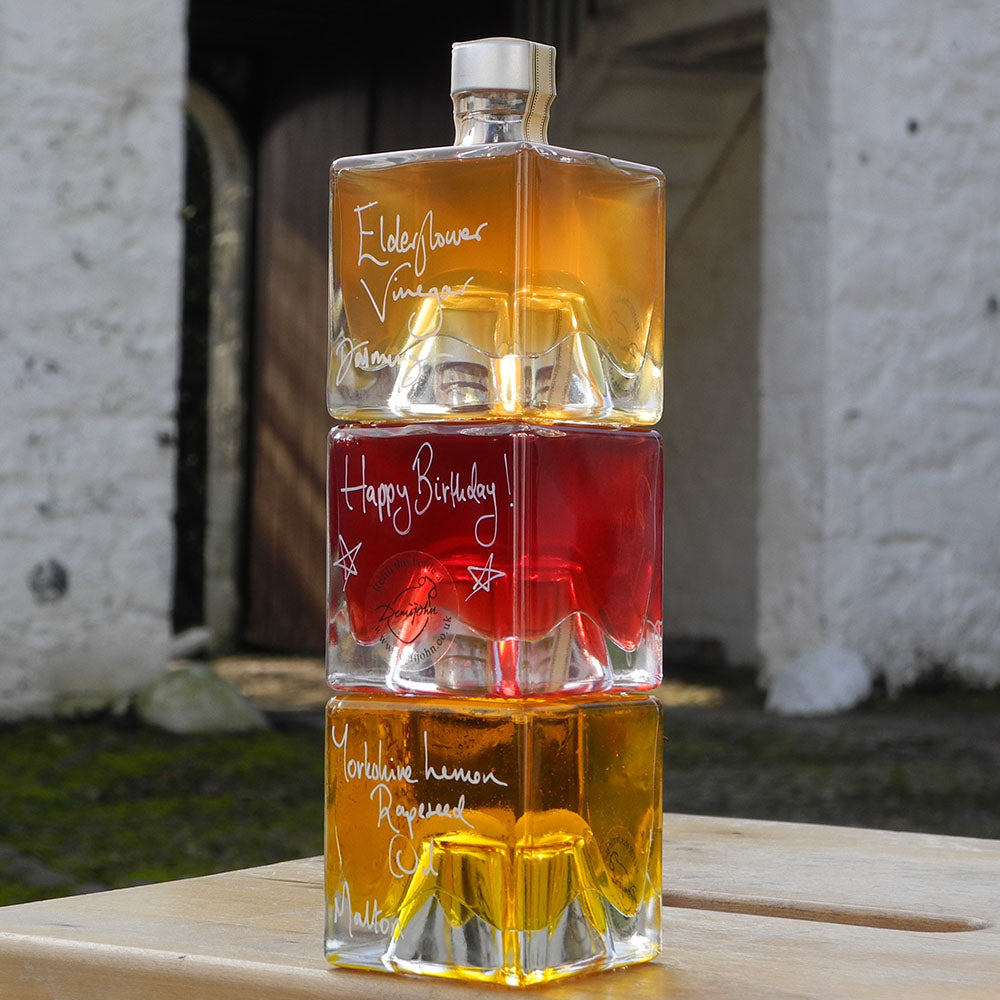 Three stacking, cube shaped, refillable 100ml glass bottles of Elderflower Vinegar, Scottish Raspberry Vinegar and Lemon infused Rapseed Oil, photographed outdoors in front of red barn doors. One bottle has a handwritten message saying Happy Birthday on it.