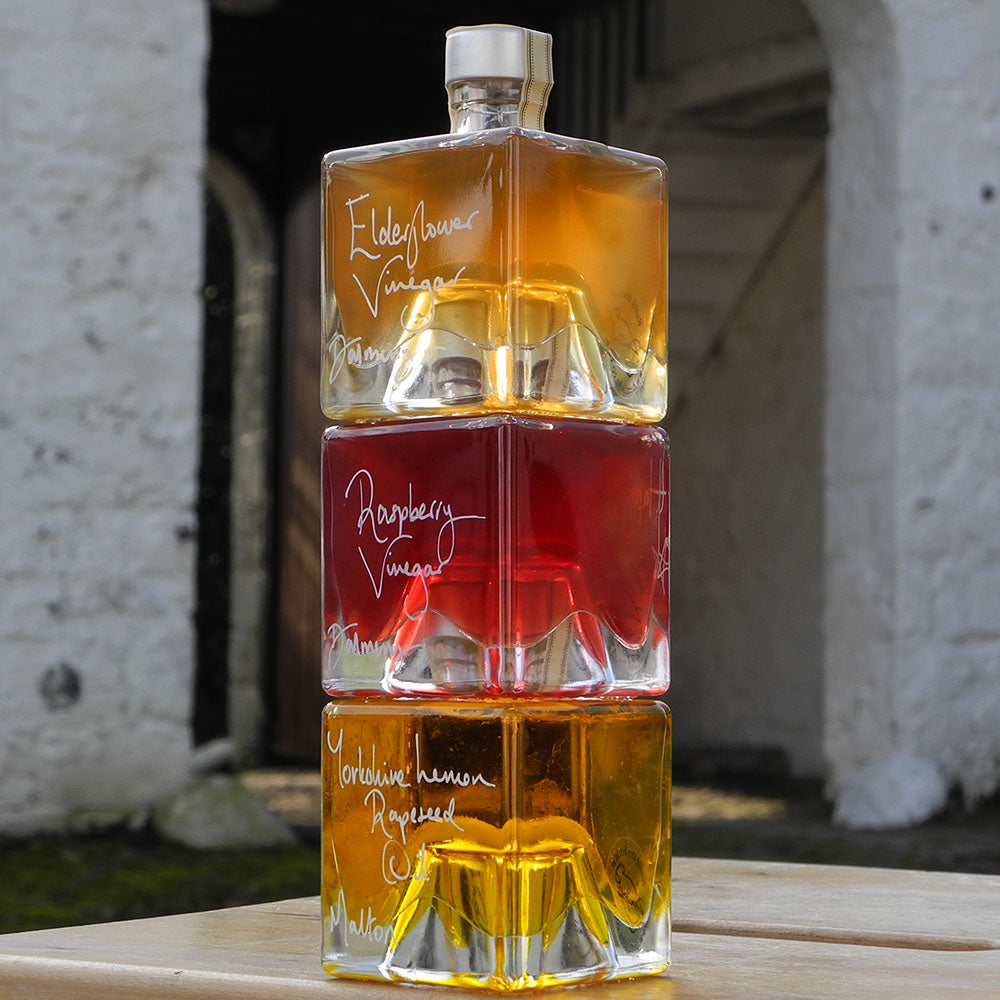 Three stacking, cube shaped, refillable 100ml glass bottles of Elderflower Vinegar, Scottish Raspberry Vinegar and Lemon infused Rapseed Oil, photographed outdoors in front of red barn doors.