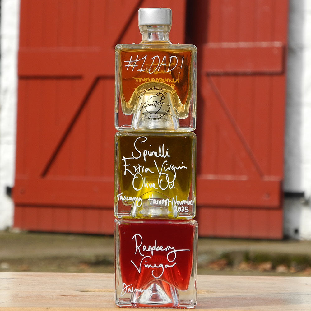 Three Mystic 100ml stacking bottles on top of one another containing Lime and Lemongrass Balsamic Vinegar, Spinelli Tuscan Extra Virgin Olive Oil and Raspberry Vinegar. The labels are handwritten in white pen in front of the red Demijohn shutters at Glenlair Steading, there is a personalised message which says Number One Dad