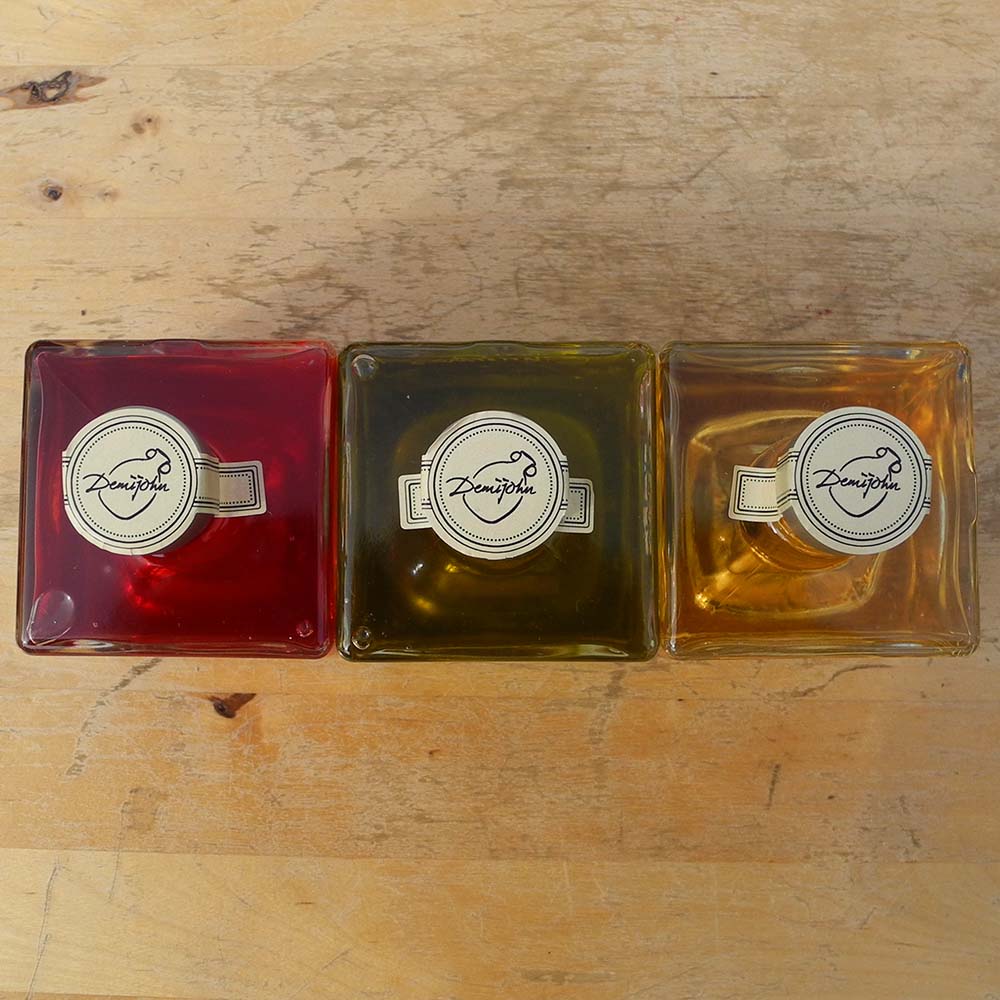 Three Mystic 100ml stacking bottles containing Lime and Lemongrass Balsamic Vinegar, Spinelli Tuscan Extra Virgin Olive Oil and Raspberry Vinegar. The labels are handwritten in white pen in front of the red Demijohn shutters at Glenlair Steading. This shot is from above and shows the Demijohn logo on the tamper proof seals.