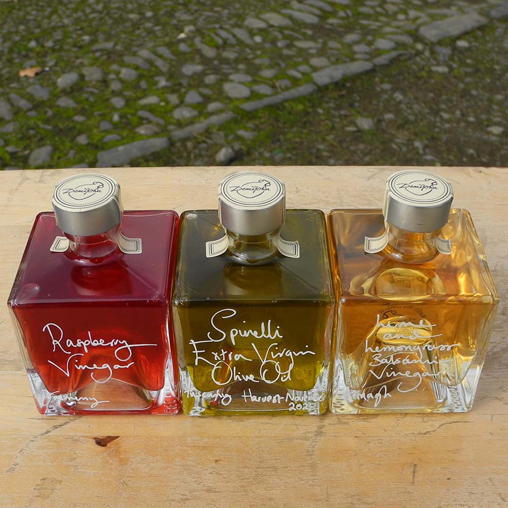 Three Mystic 100ml stacking bottles containing Lime and Lemongrass Balsamic Vinegar, Spinelli Tuscan Extra Virgin Olive Oil and Raspberry Vinegar. The labels are handwritten in white pen in front of the red Demijohn shutters at Glenlair Steading. 
