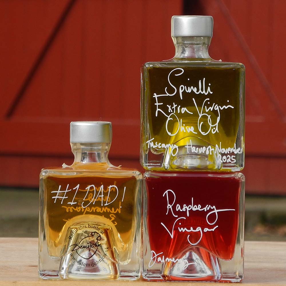Three Mystic 100ml stacking bottles on top of one another containing Lime and Lemongrass Balsamic Vinegar, Spinelli Tuscan Extra Virgin Olive Oil and Raspberry Vinegar. The labels are handwritten in white pen in front of the red Demijohn shutters at Glenlair Steading, there is a personalised message which says number one dad.