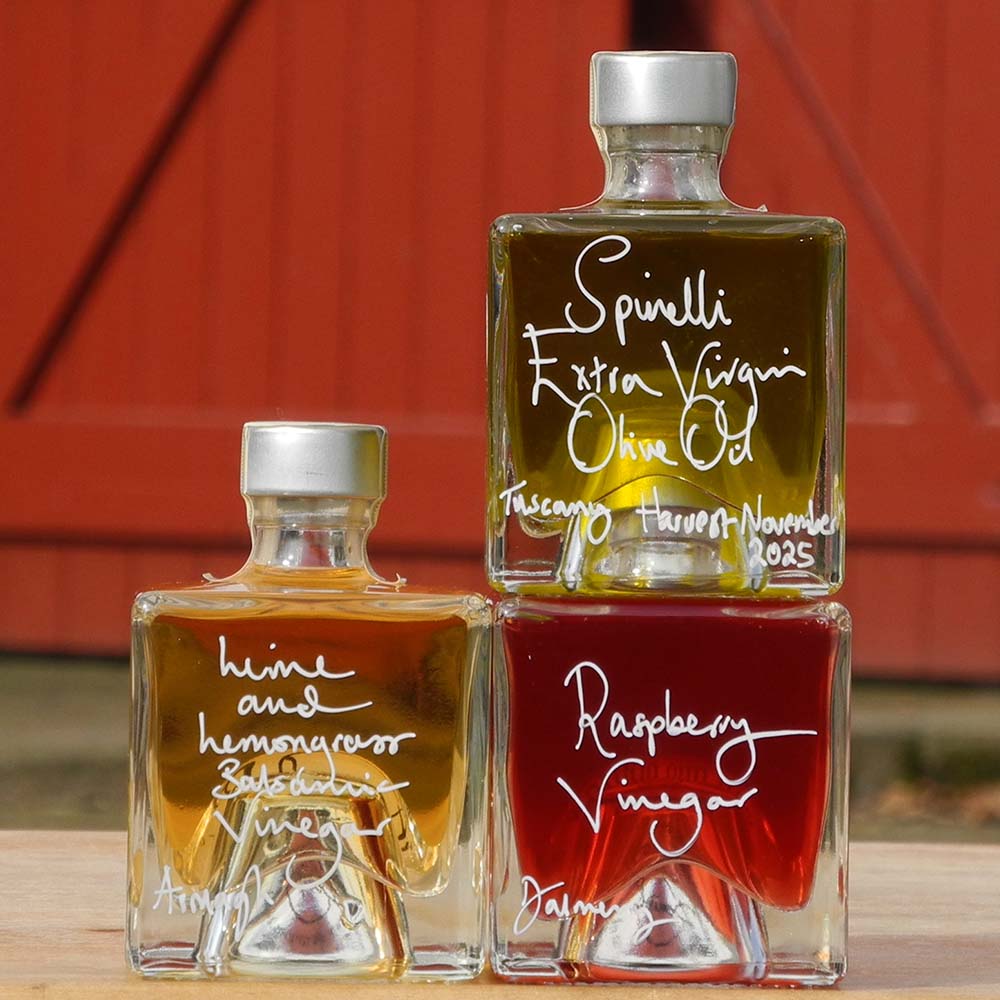 Three Mystic 100ml stacking bottles on top of one another containing Lime and Lemongrass Balsamic Vinegar, Spinelli Tuscan Extra Virgin Olive Oil and Raspberry Vinegar. The labels are handwritten in white pen in front of the red Demijohn shutters at Glenlair Steading.