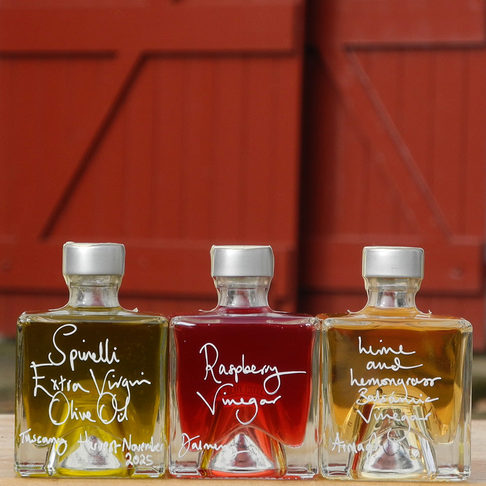 Three Mystic 100ml stacking bottles on top of one another containing Lime and Lemongrass Balsamic Vinegar, Spinelli Tuscan Extra Virgin Olive Oil and Raspberry Vinegar. The labels are handwritten in white pen in front of the red Demijohn shutters at Glenlair Steading.