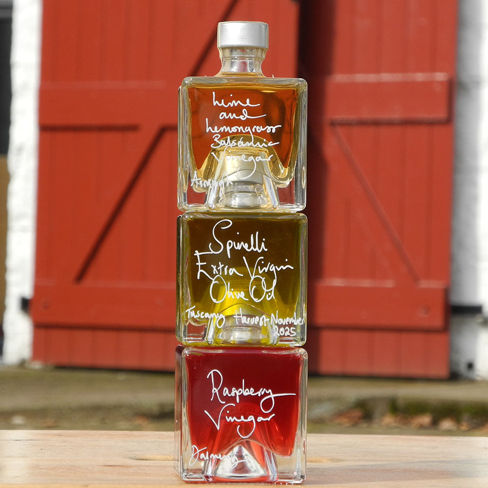 Three Mystic 100ml stacking bottles on top of one another containing Lime and Lemongrass Balsamic Vinegar, Spinelli Tuscan Extra Virgin Olive Oil and Raspberry Vinegar. The labels are handwritten in white pen in front of the red Demijohn shutters at Glenlair Steading.