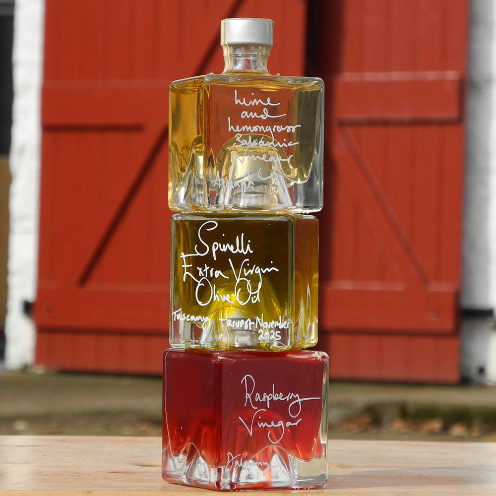 Three Mystic 100ml stacking bottles on top of one another containing Lime and Lemongrass Balsamic Vinegar, Spinelli Tuscan Extra Virgin Olive Oil and Raspberry Vinegar. The labels are handwritten in white pen in front of the red Demijohn shutters at Glenlair Steading.