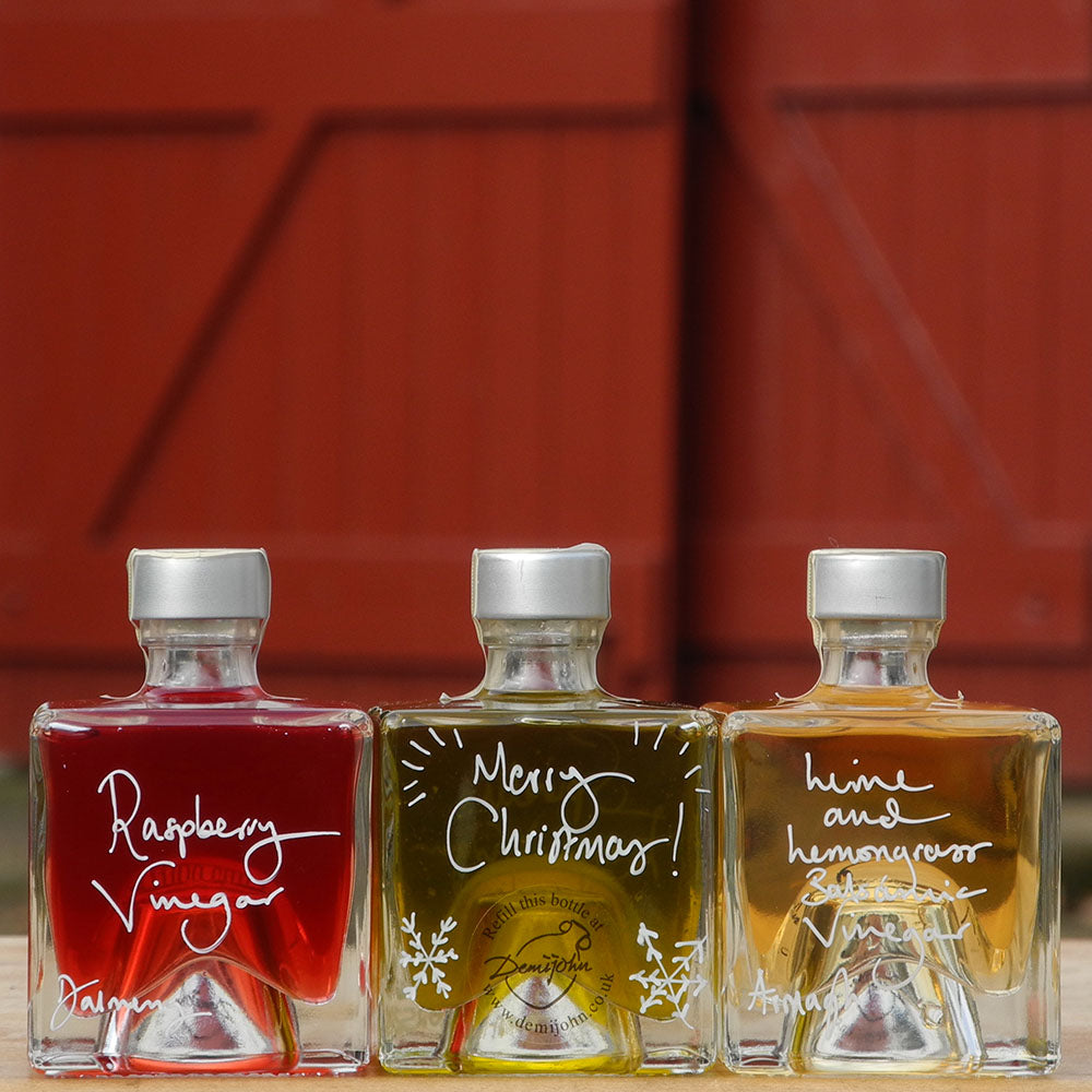 Three Mystic 100ml stacking bottles containing Lime and Lemongrass Balsamic Vinegar, Spinelli Tuscan Extra Virgin Olive Oil and Raspberry Vinegar. The labels are handwritten in white pen in front of the red Demijohn shutters at Glenlair Steading. there is a personalised message which says Merry Christmas